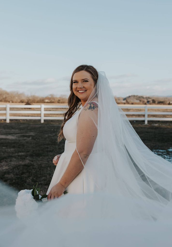 Bride smiling over her shoulder in a white wedding gown with a long flowing veil, holding a bouquet of white flowers in front of a countryside fence.