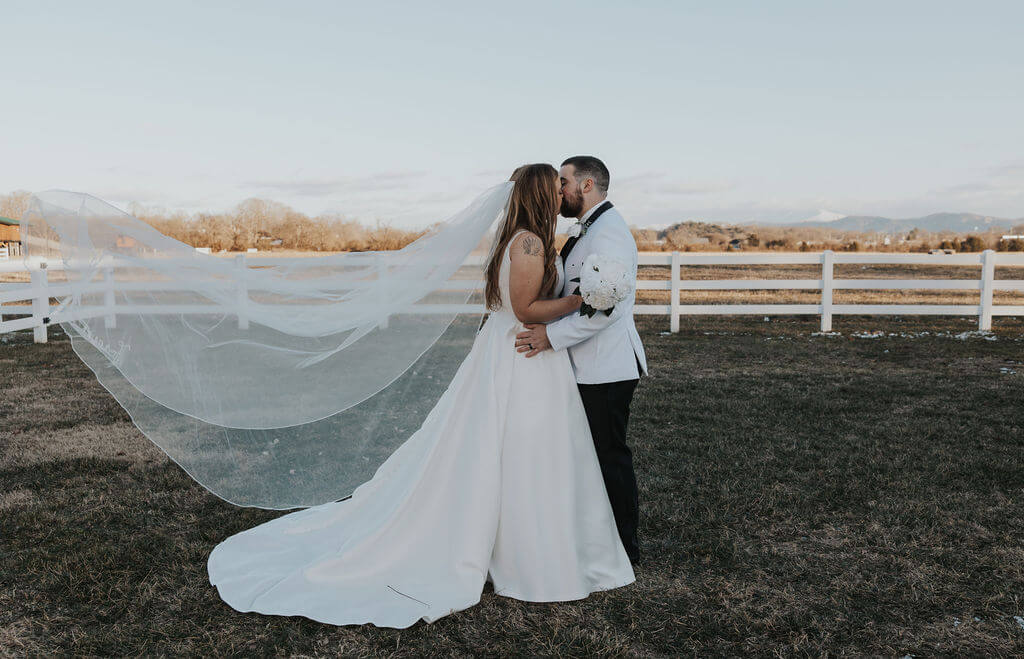 Bride and groom kissing outdoors in a grassy field, with the bride's long wedding veil blowing dramatically in the wind against a white fence and distant mountains.
