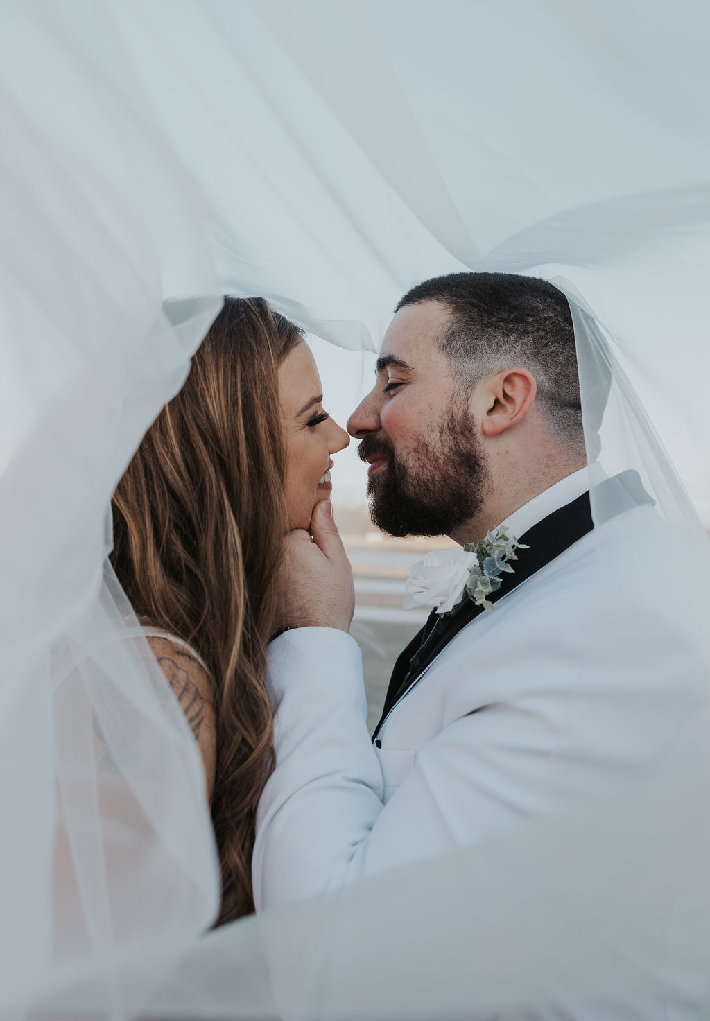 Close-up of a bride and groom smiling at each other beneath her sheer wedding veil, with the groom gently holding the bride's chin.