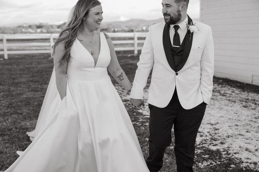 Black-and-white candid photo of a bride and groom holding hands and smiling at each other while walking outside their wedding venue, with a white fence in the background.
