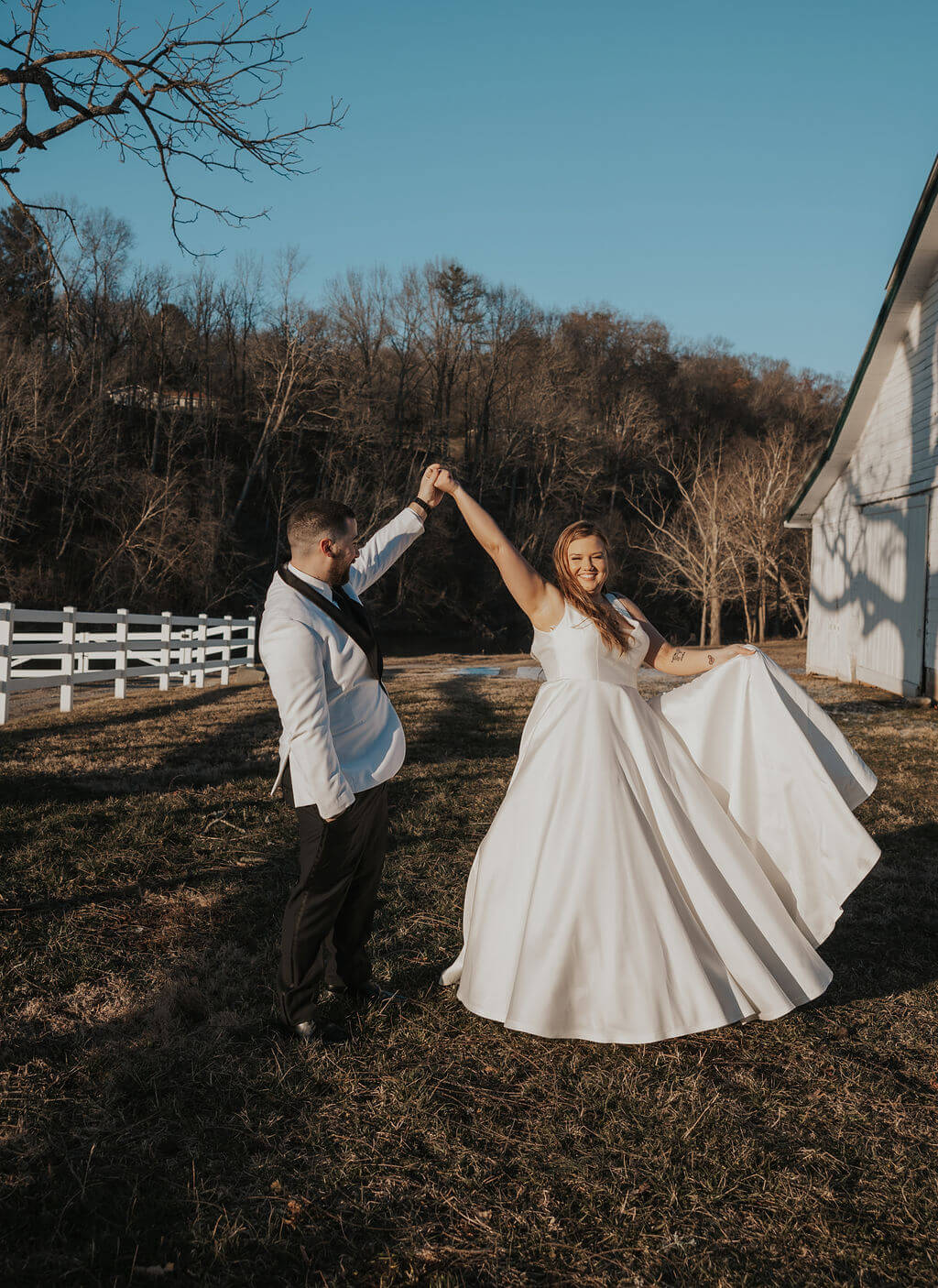 Groom twirling his bride in her flowing white wedding dress outside a rustic white barn, with a wooden fence and bare trees in the background.