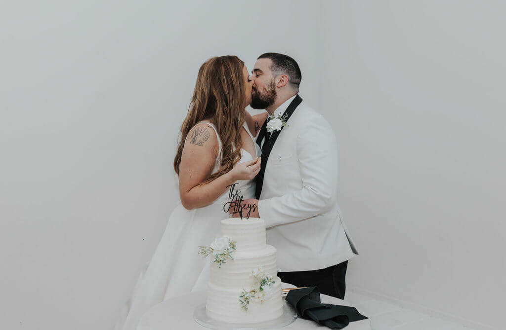 Bride and groom sharing a kiss behind a three-tier white wedding cake decorated with delicate white flowers and a personalized cake topper.
