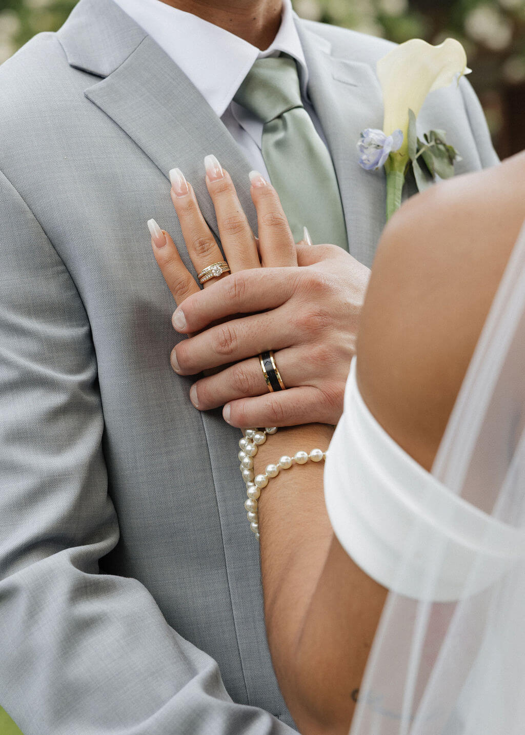 Close-up of a bride’s hand resting on the groom’s gray suit, showing their wedding rings and her pearl bracelet.