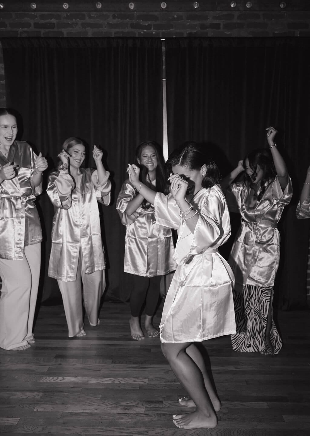 Black-and-white candid of the bride dancing in a satin robe with her bridesmaids while getting ready for the wedding.