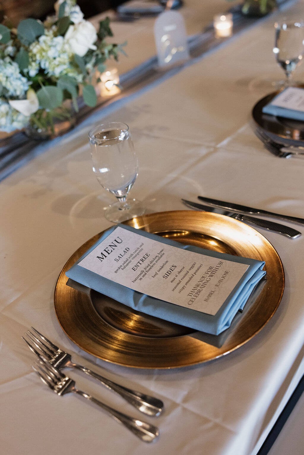 Wedding reception place setting with a gold charger, blue napkin, menu card, glassware, and floral centerpiece.