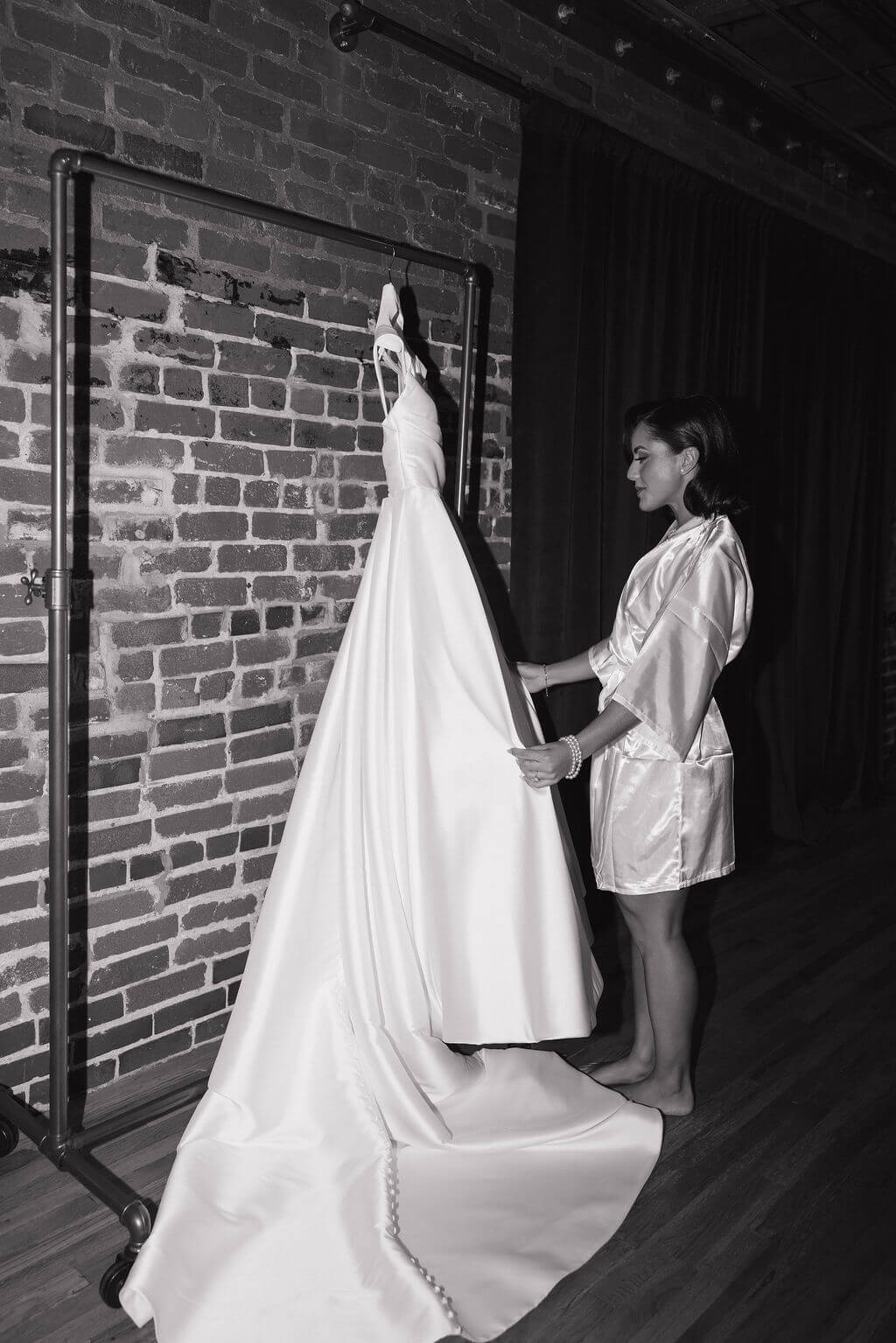 Black-and-white photo of the bride standing beside her wedding gown hanging against an exposed brick wall.