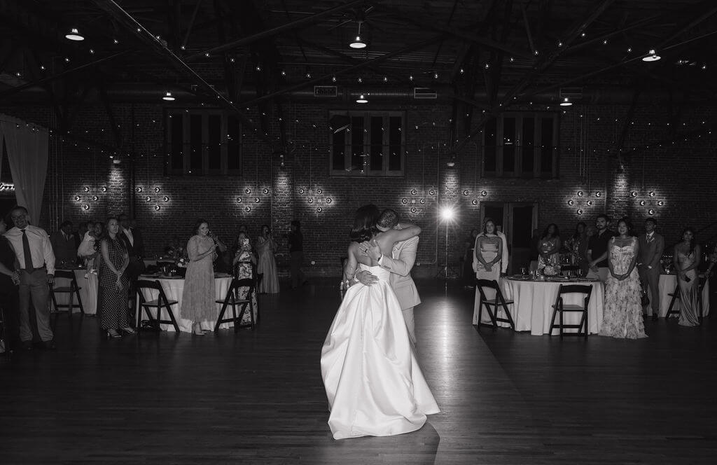 Black-and-white photo of the bride and groom sharing their first dance inside The Mill and Mine as wedding guests watch.