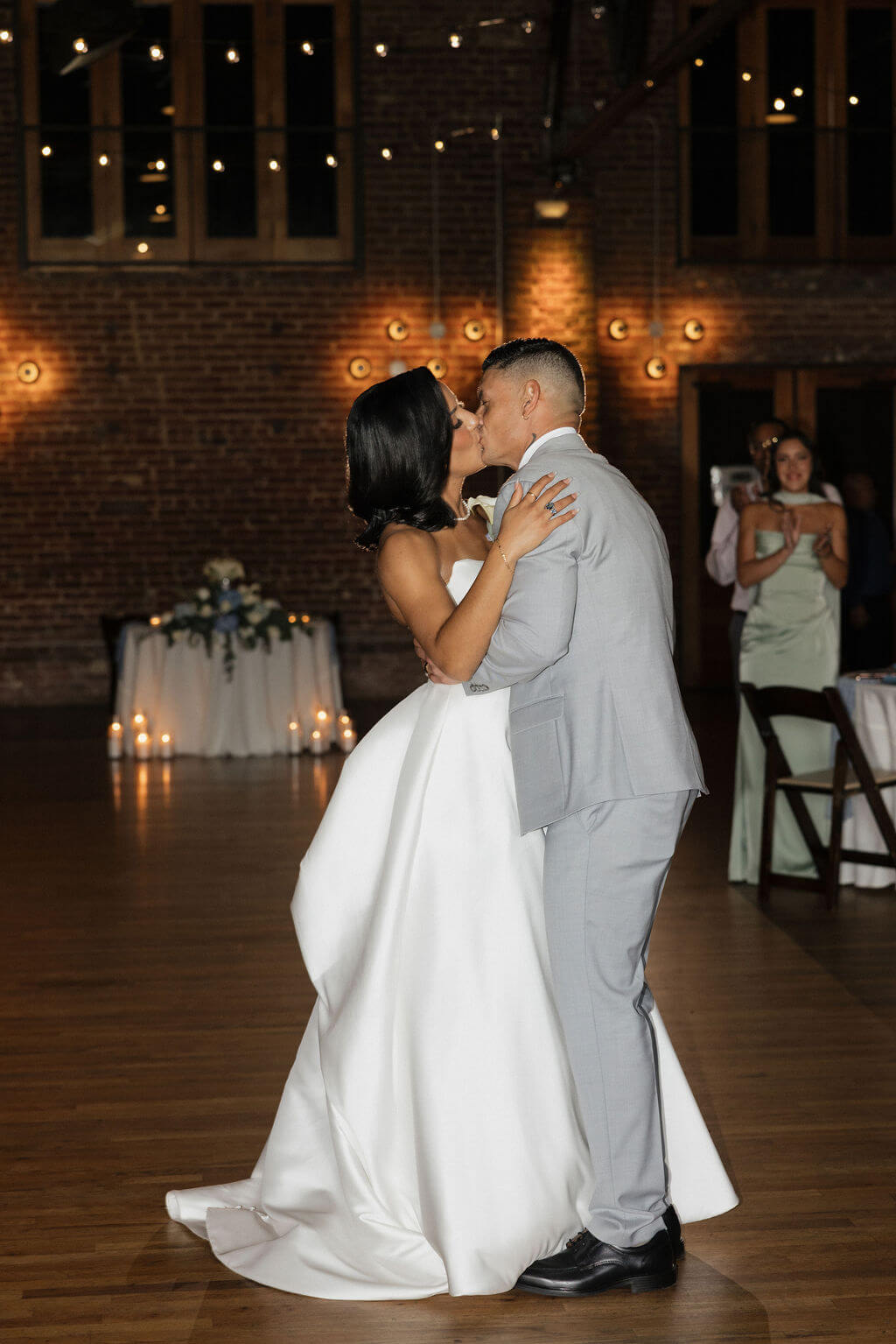 Bride and groom kissing during their first dance with warm lights glowing against The Mill and Mine’s brick walls.