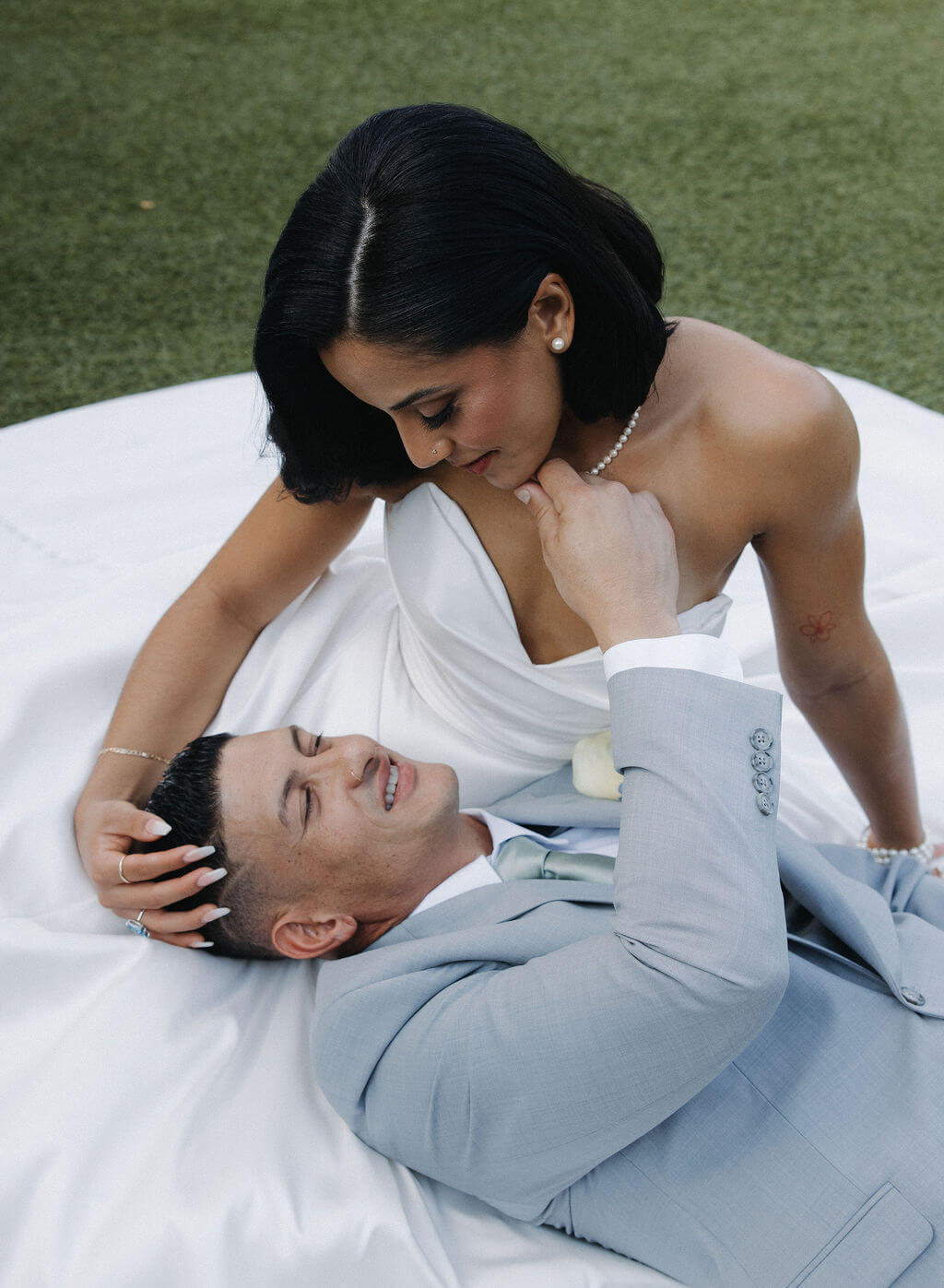 Bride sitting on the grass while the groom rests his head in her lap during relaxed wedding portraits.