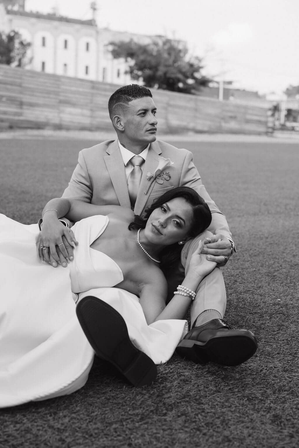 Black-and-white portrait of the bride reclining against the groom as they sit together on the grass.