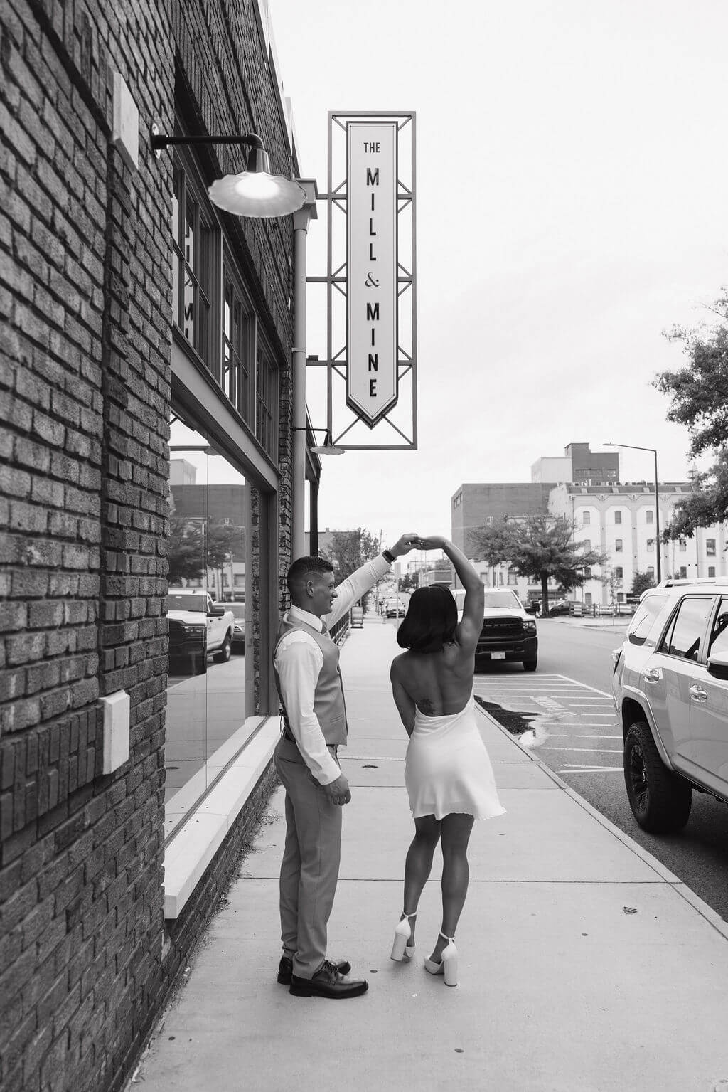 Black-and-white photo of the groom twirling the bride in her short white reception dress on the sidewalk outside The Mill and Mine.