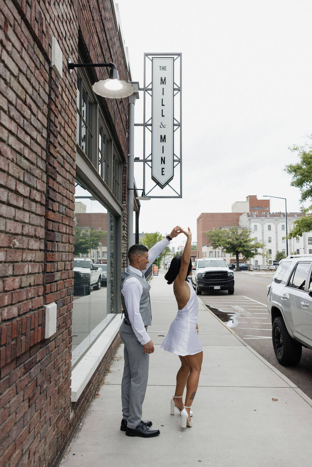 Bride in a short white reception dress twirling with the groom on the sidewalk beneath The Mill and Mine sign.