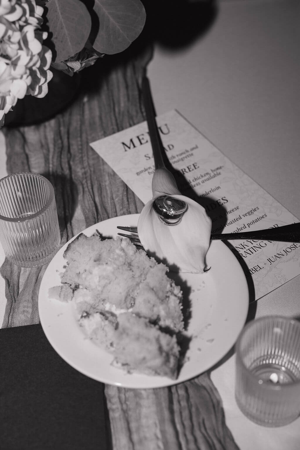 Black-and-white detail of a slice of wedding cake beside the reception menu and glassware.