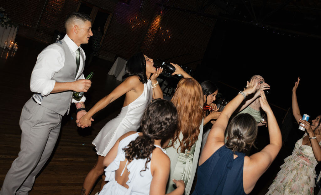 Bride drinking directly from a champagne bottle while dancing with the groom and wedding guests at the reception.