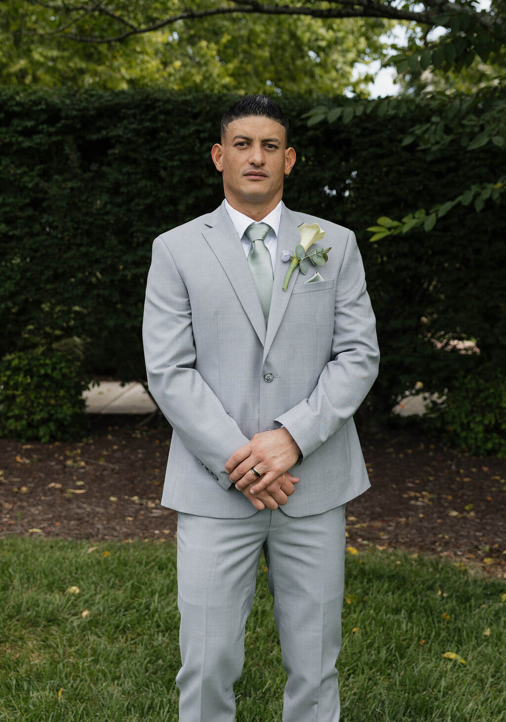 Groom posing outdoors in a light gray suit with a pale green tie and boutonniere.