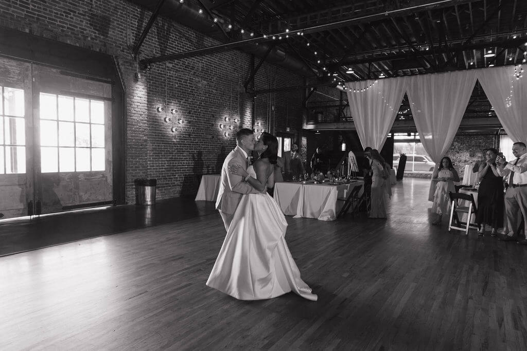 Black-and-white wide shot of the bride and groom dancing inside The Mill and Mine with large windows, exposed brick walls, and guests surrounding them.