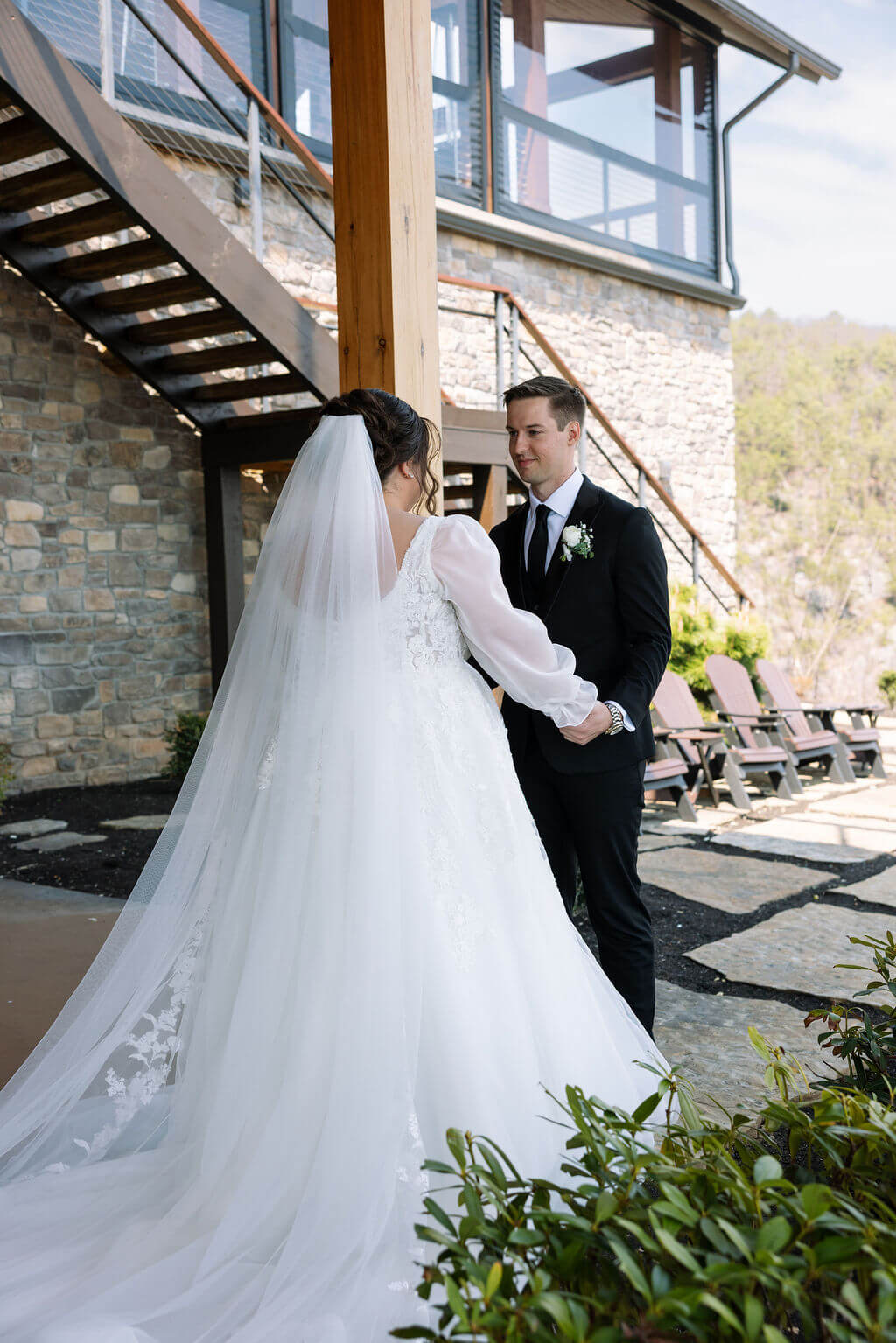 Bride and groom face each other beside a stone staircase outside a rustic wedding venue.