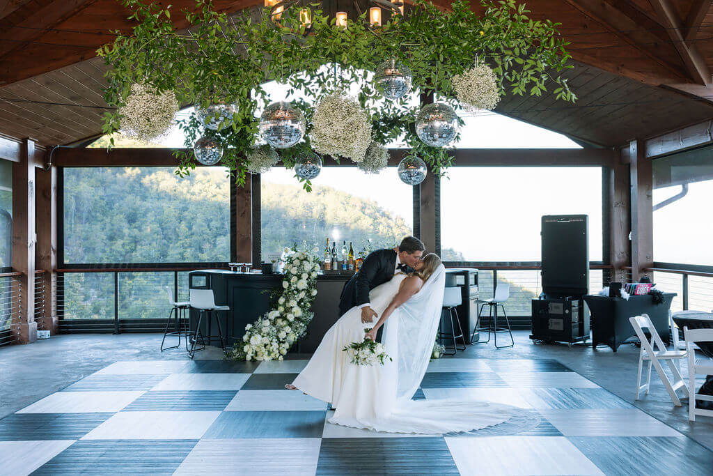 Groom dips the bride for a kiss on a black-and-white checkered dance floor beneath hanging greenery and disco balls.
