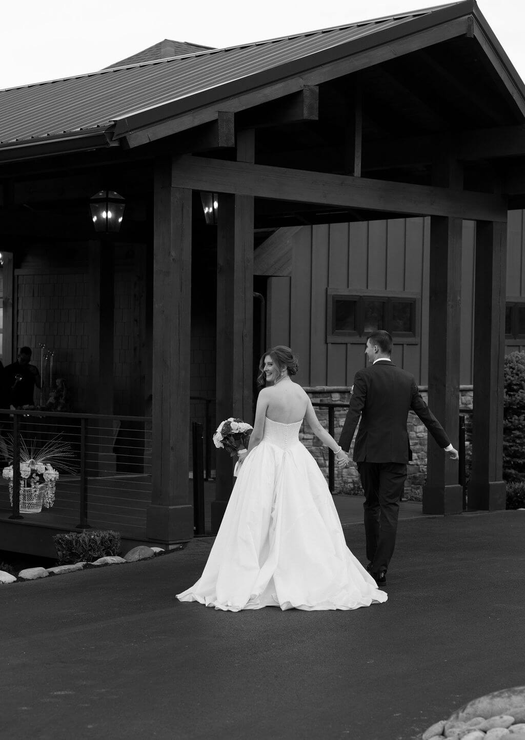 Black-and-white photograph of newlyweds walking hand in hand outside a rustic wedding venue.