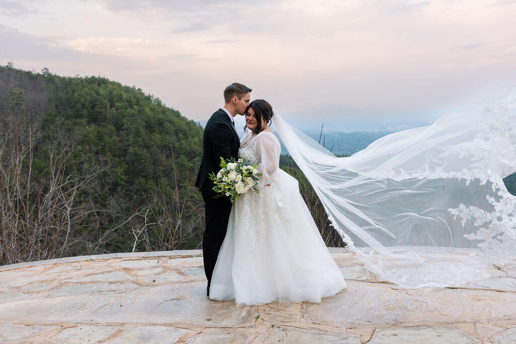 Bride and groom stand together on a stone overlook as her long veil blows dramatically in the wind.