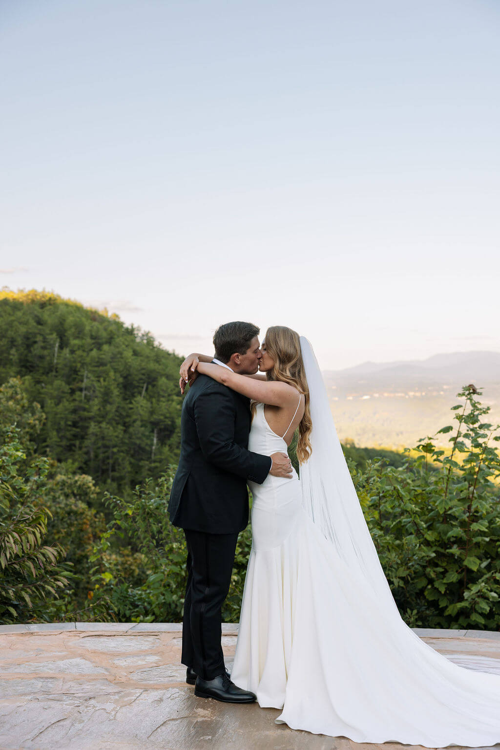 Bride and groom kiss on a stone overlook surrounded by mountains and lush green hills.