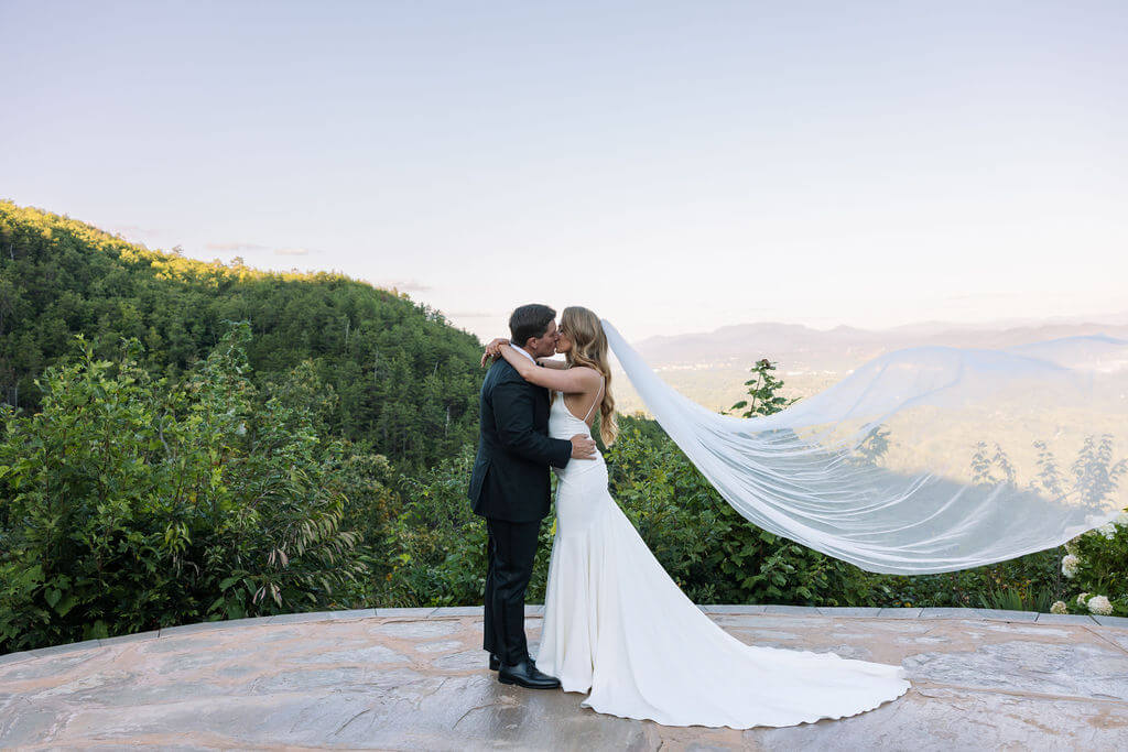 Bride and groom kiss on a stone overlook as her long veil flows behind them against a mountain backdrop.