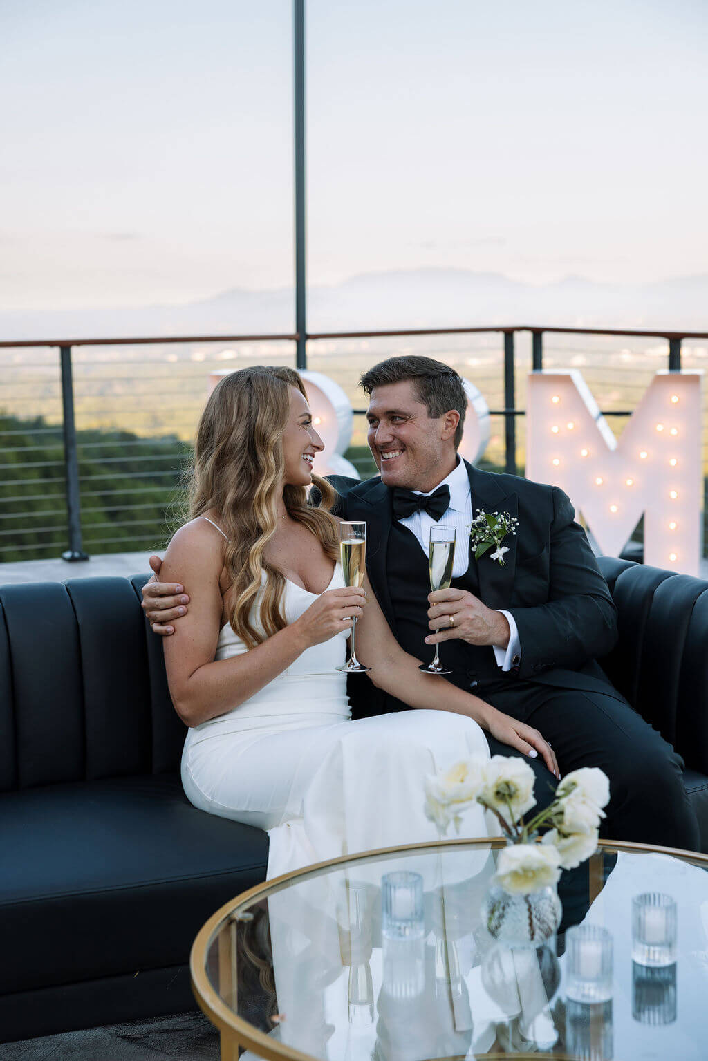 Bride and groom sit together on a black couch during their wedding reception, smiling at each other.