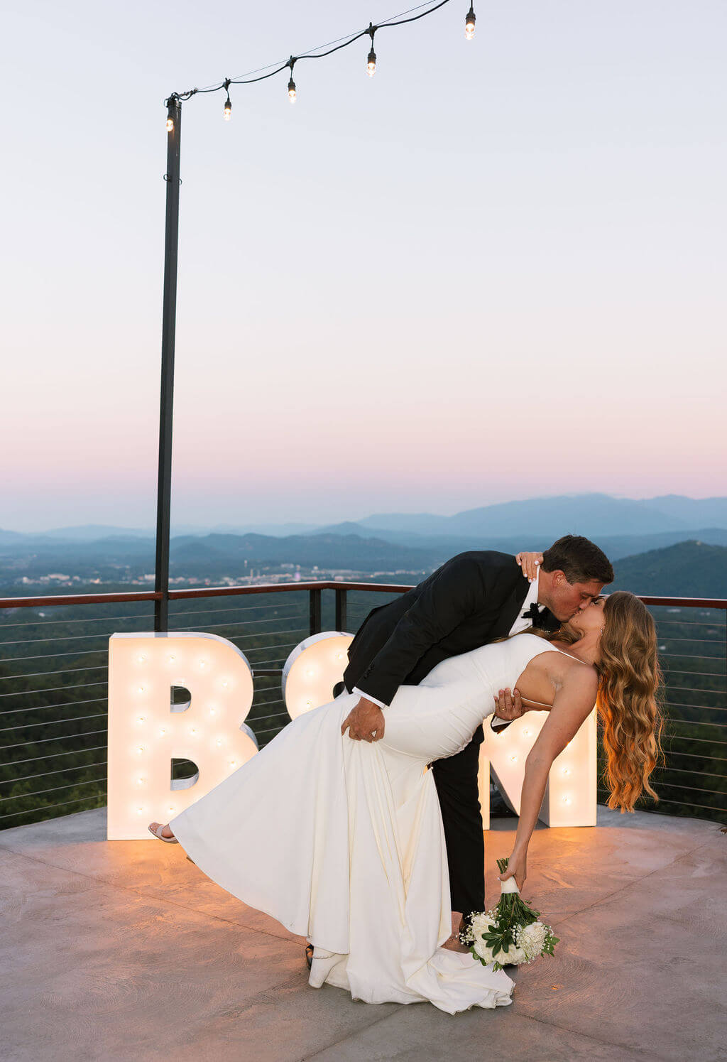 Groom dips the bride for a kiss beside illuminated marquee letters on an outdoor wedding terrace at sunset.