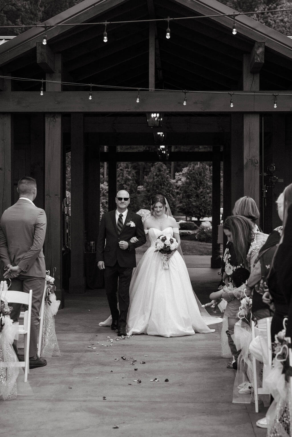 Black-and-white photograph of a bride walking down the aisle with an escort during an outdoor wedding ceremony.