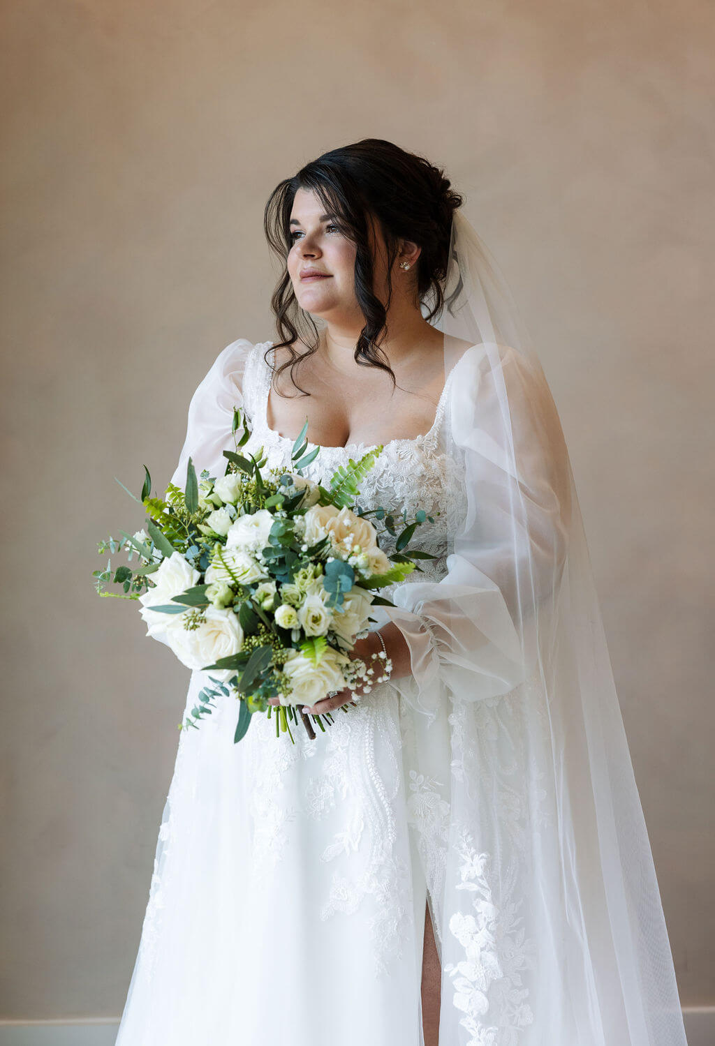 Bride holds a bouquet of white flowers and greenery while standing beside a window in her wedding dress.