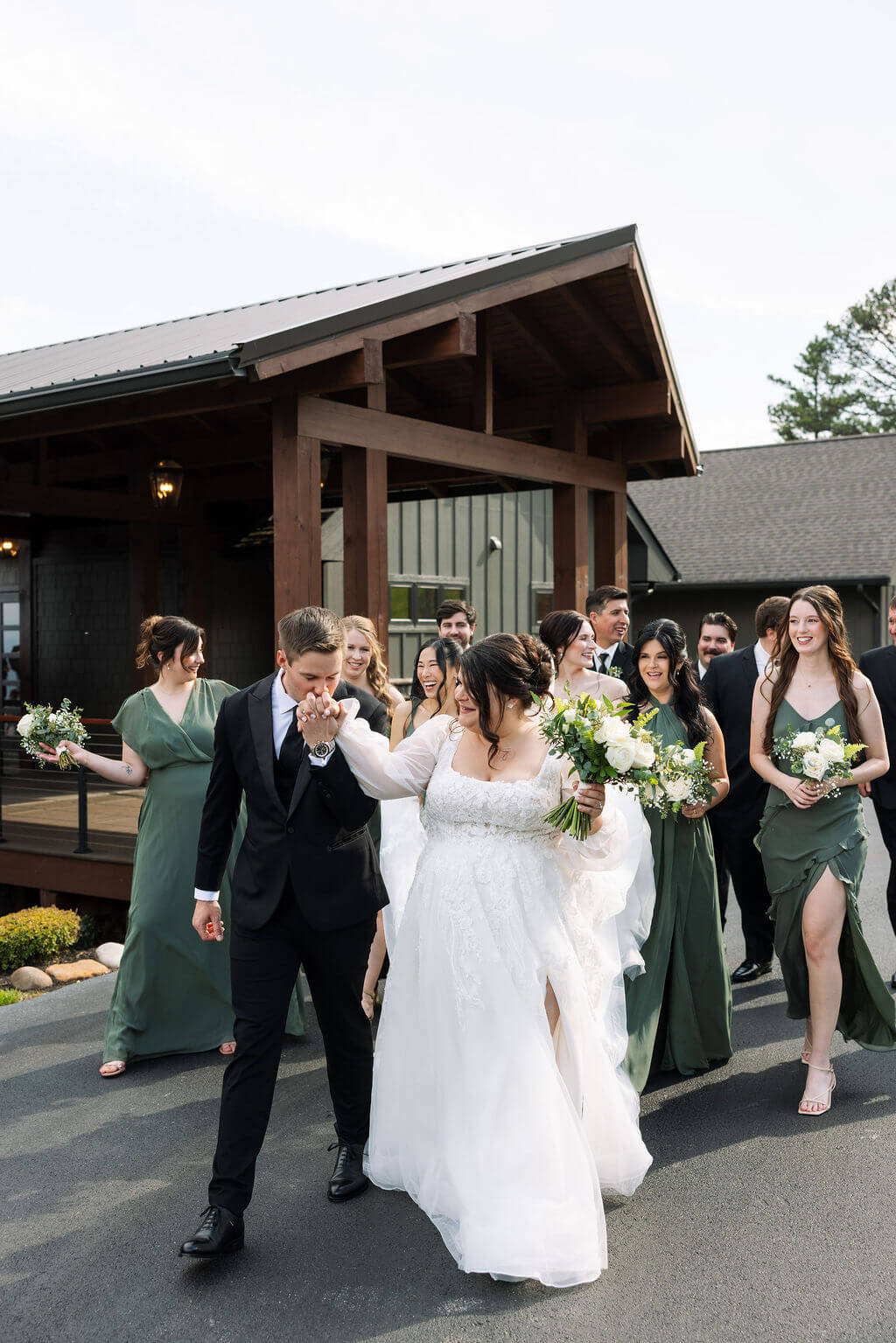 Bride in a flowing white wedding dress walks alongside the groom and bridesmaids in green dresses outside a mountain wedding venue.