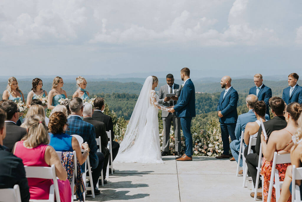Bride and groom exchange vows during an outdoor wedding ceremony with mountain views and guests seated on either side of the aisle.