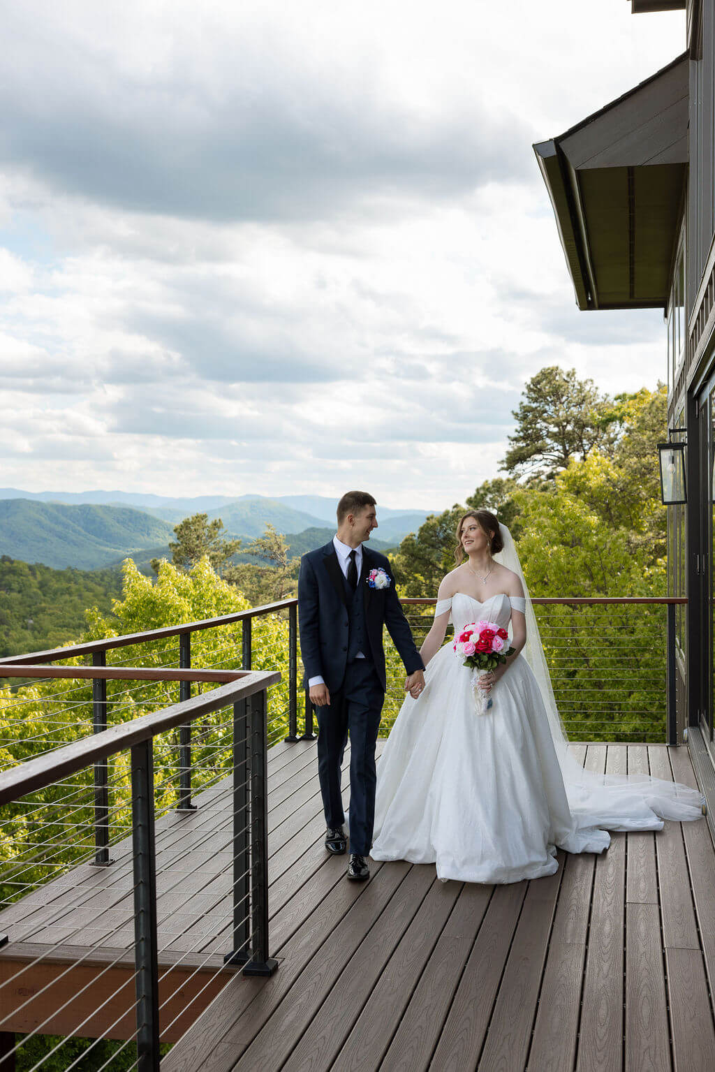 Bride and groom walk hand in hand along a wooden balcony with mountain views behind them.