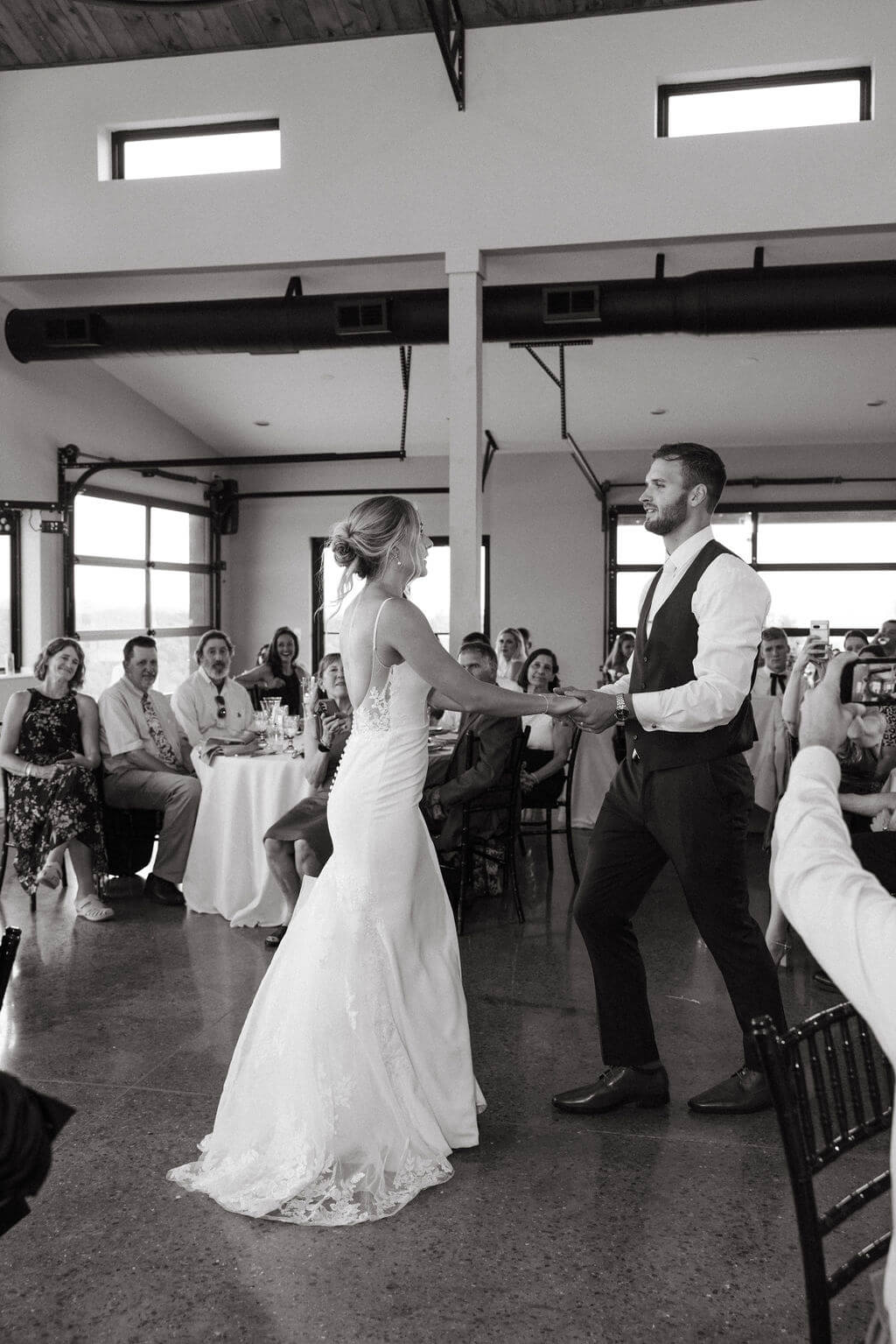 Black-and-white photograph of newlyweds dancing inside a wedding reception venue as guests watch.