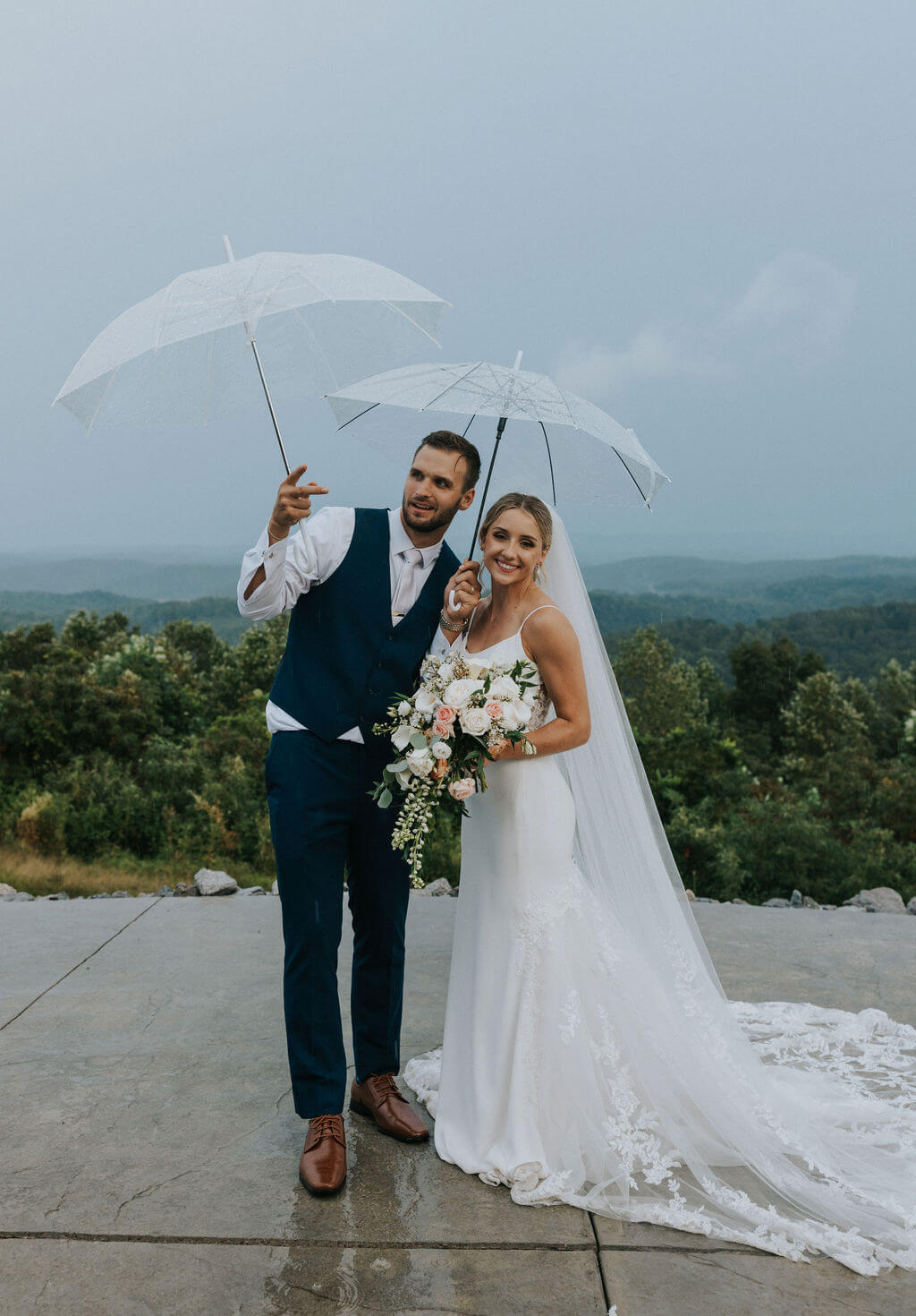 Bride and groom smile beneath a clear umbrella with mountain scenery in the background.