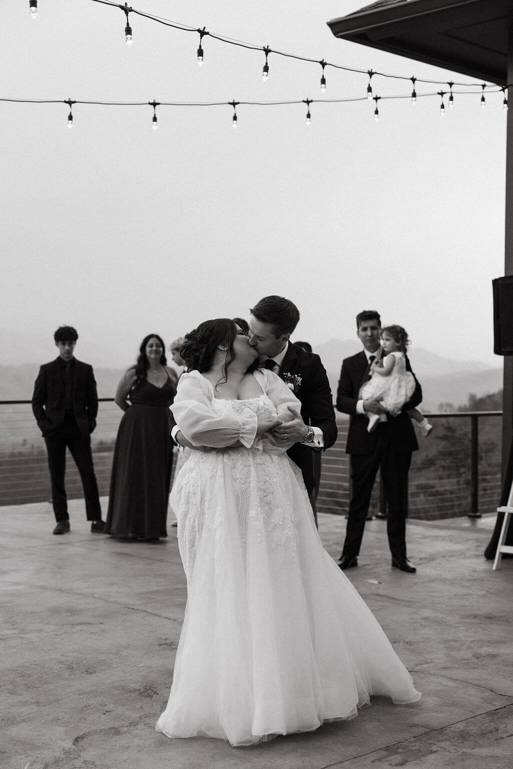 Black-and-white photograph of a bride and groom dancing outdoors beneath string lights as wedding guests watch.