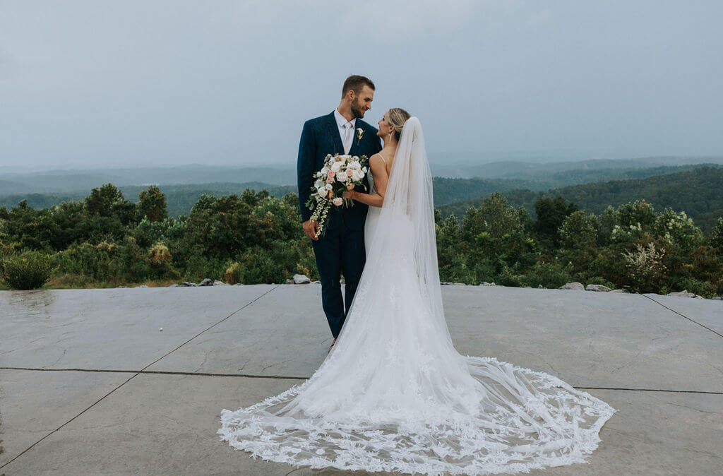 Bride and groom embrace on a mountain overlook with rolling green hills stretching into the distance.