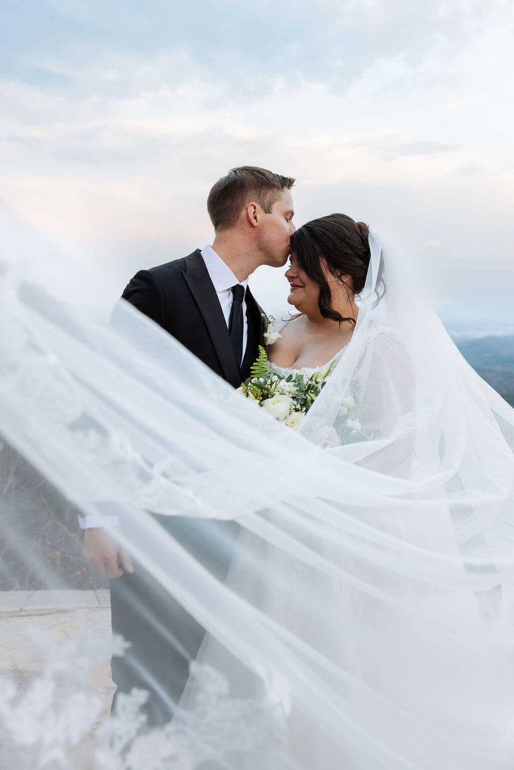 Groom kisses the bride's forehead while her flowing veil frames them against the mountain landscape.