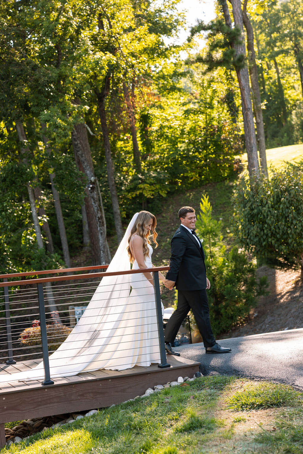 Newlyweds walk hand in hand across a wooden bridge surrounded by trees and greenery.
