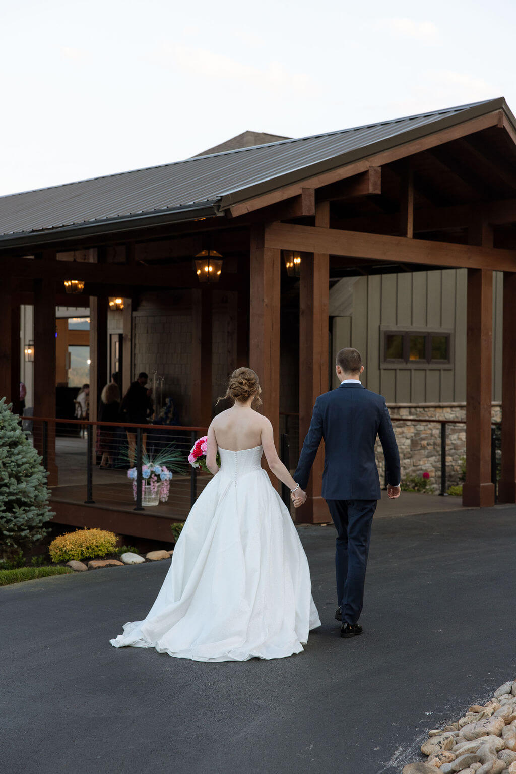 Newlyweds walk hand in hand toward a modern rustic wedding venue with wooden beams and a covered porch.